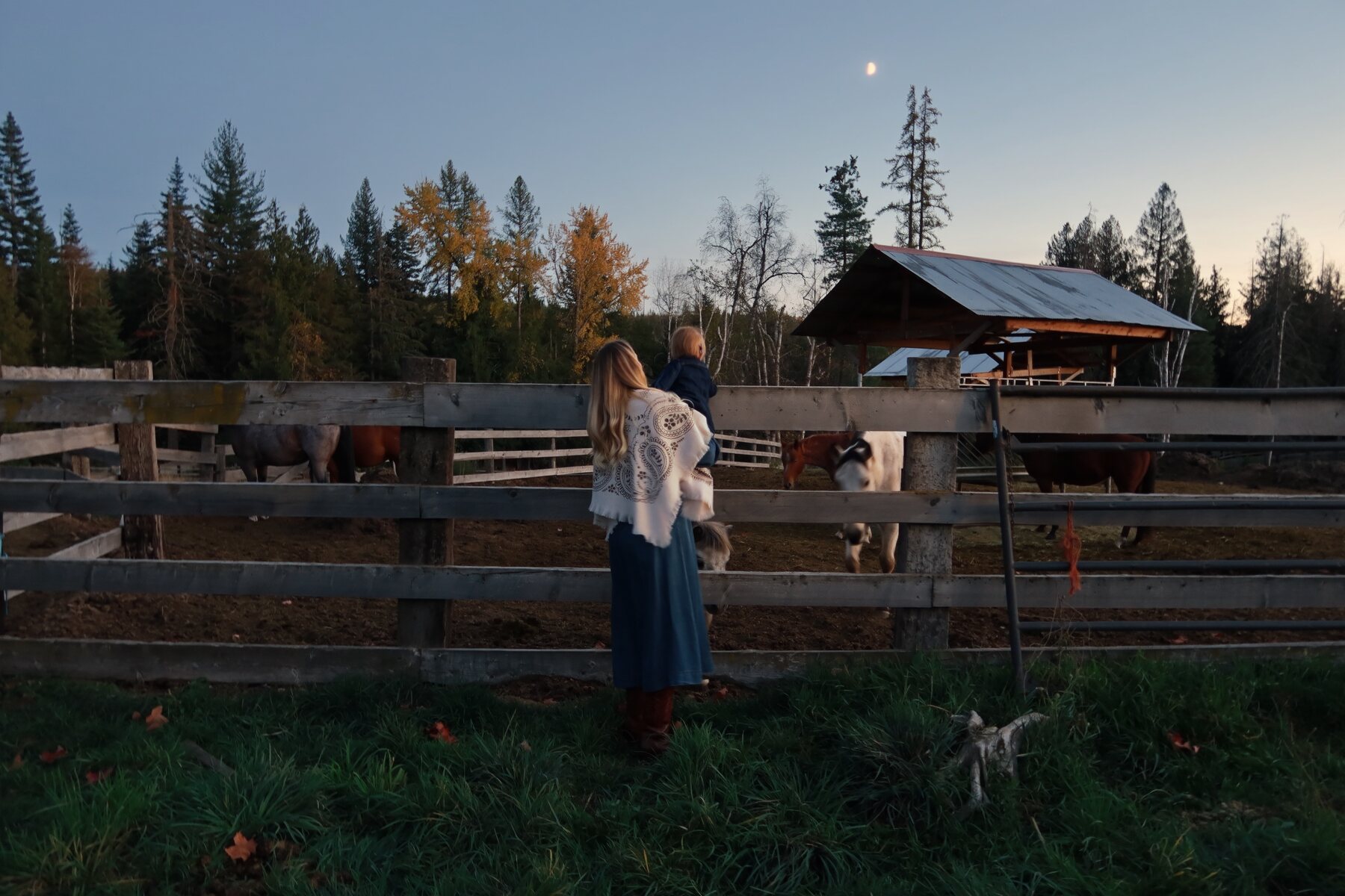 Kiana holding our daughter at the corral fence with the horses at dusk under the moon